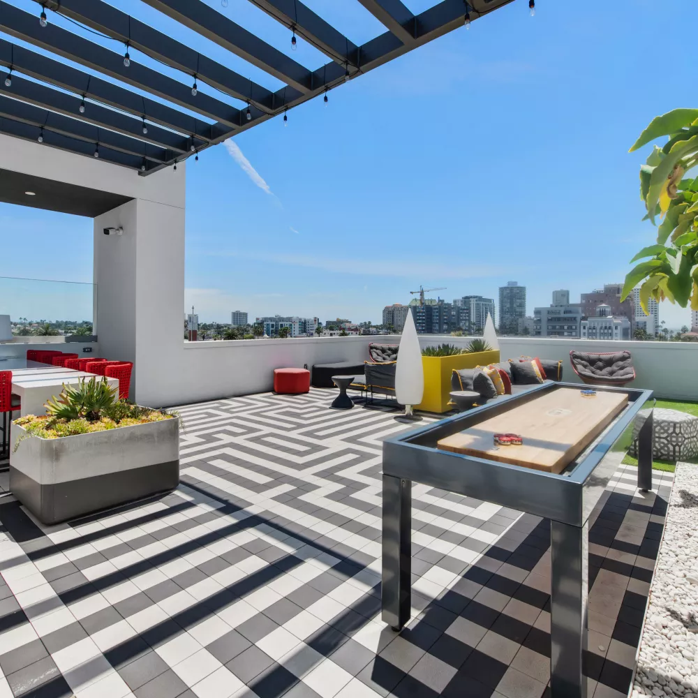Spacious rooftop patio with black and white geometric tile, pergola, lounge seating, shuffleboard, and city skyline.