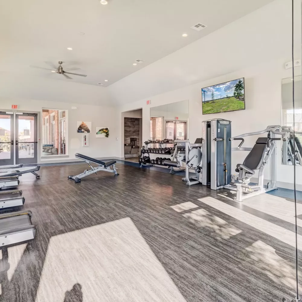 Bright, spacious gym with treadmills, weight benches, free weights, and various strength training machines under a vaulted ceiling.