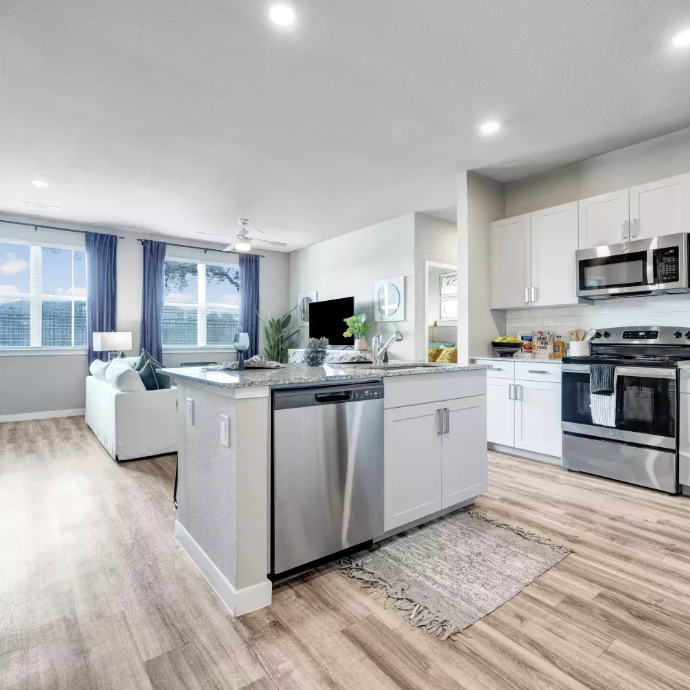 Bright, open apartment living room and kitchen with white cabinets, stainless appliances, and a white sofa by blue curtains.