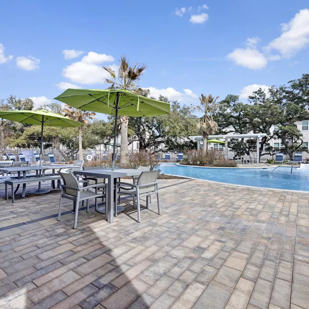 Paved patio area with outdoor dining sets, green umbrellas, and a swimming pool under a blue sky.