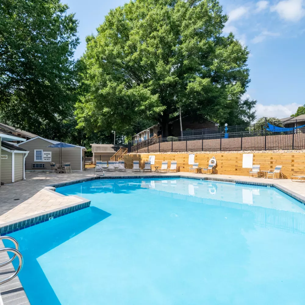 Sunny outdoor swimming pool area with a light blue pool, a tan clubhouse, and several lounge chairs.