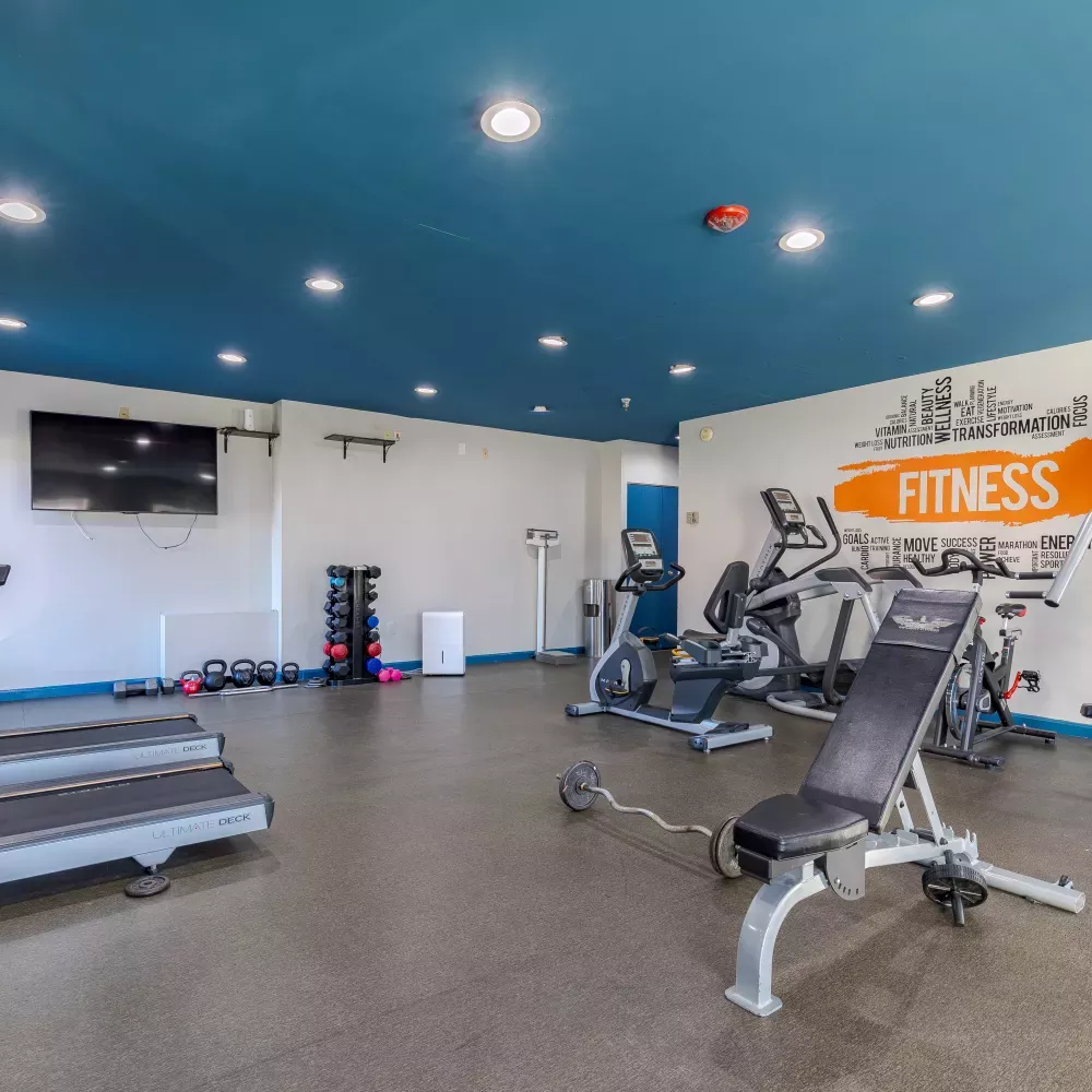 Modern gym with blue ceiling, white walls, treadmills, bikes, weight bench, free weights, and an orange "FITNESS" graphic.