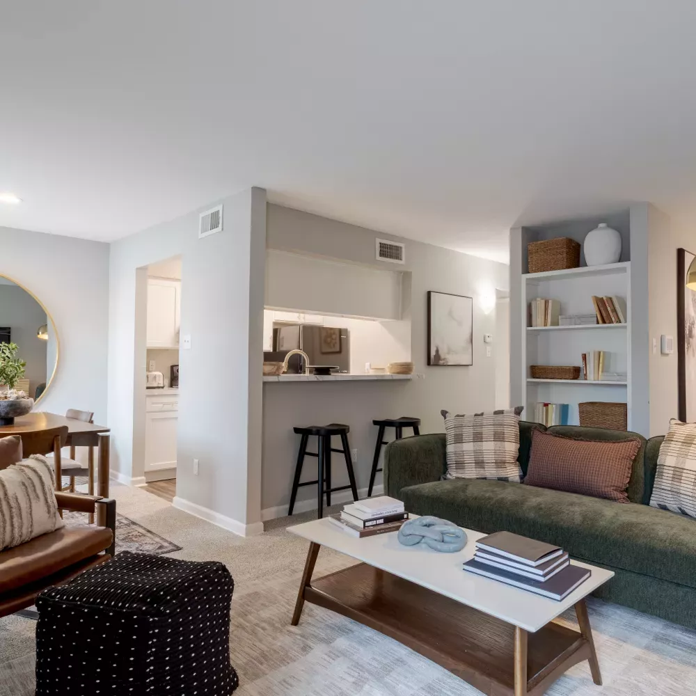 Modern living room with a green sofa, coffee table, bar, and a dining area visible through a large round mirror.