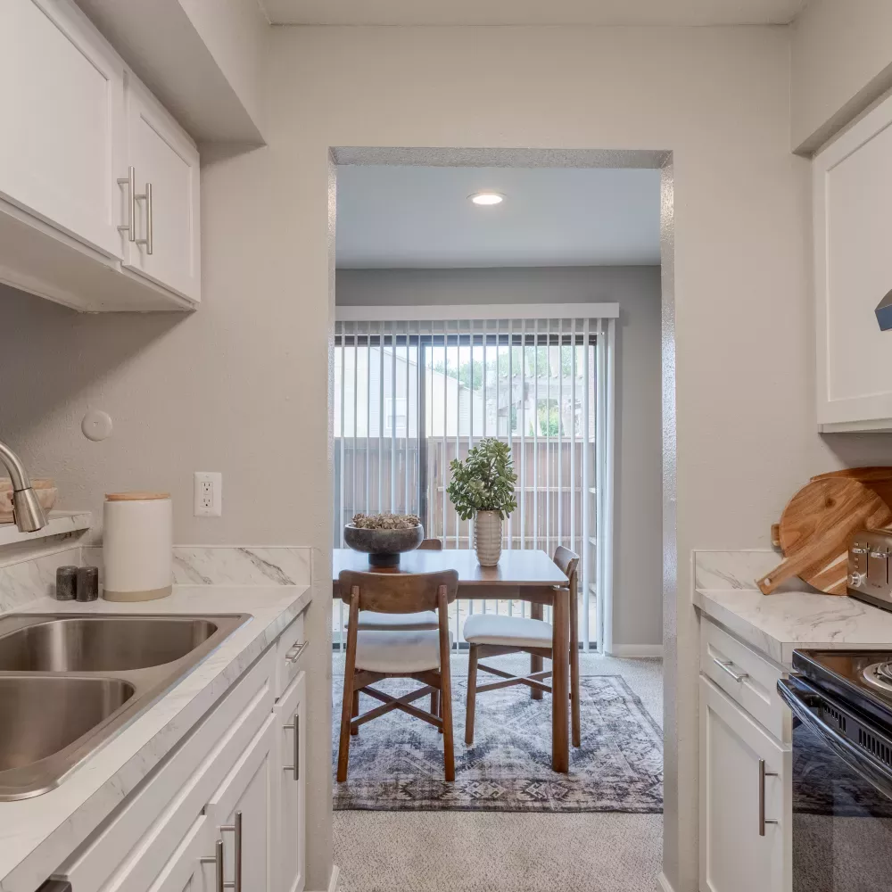 Photograph of a modern white kitchen with marble counters, double sink, and stove, looking into a dining area with a table and glass door.