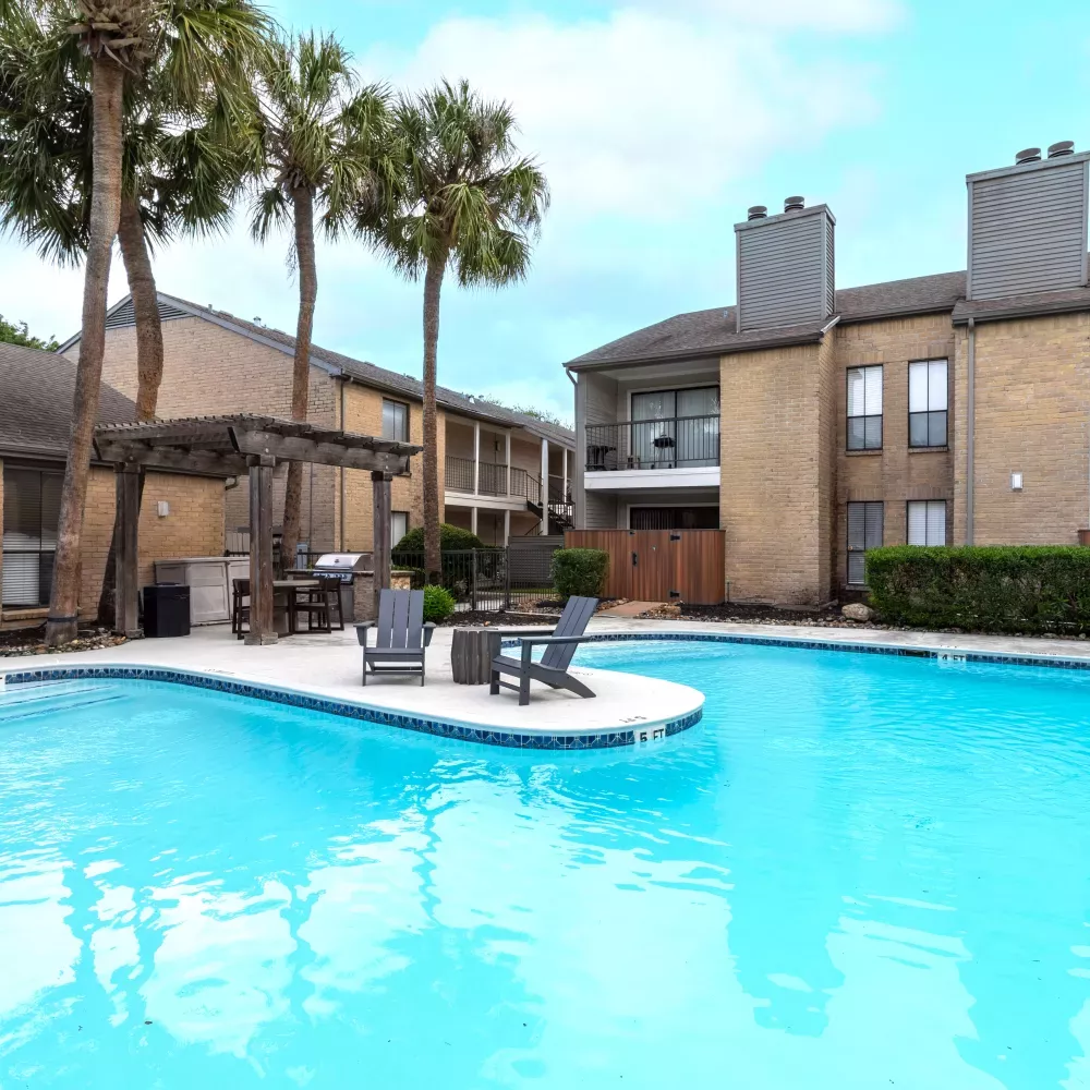 Outdoor swimming pool with light blue water, surrounded by apartment buildings, palm trees, and a shaded seating area.