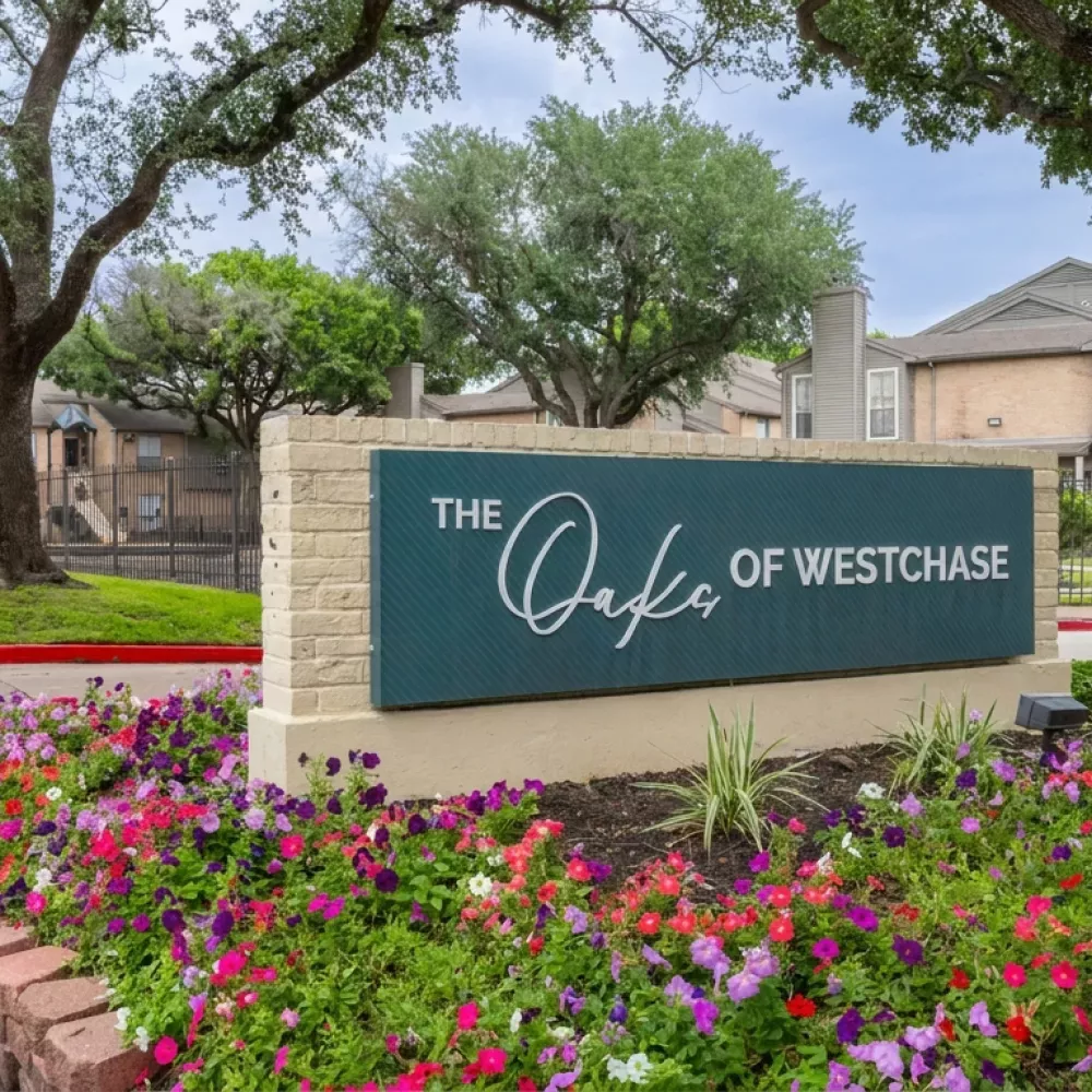 Green sign reading "The Oaks of Westchase" at an apartment complex entrance, bordered by colorful flowers and trees.