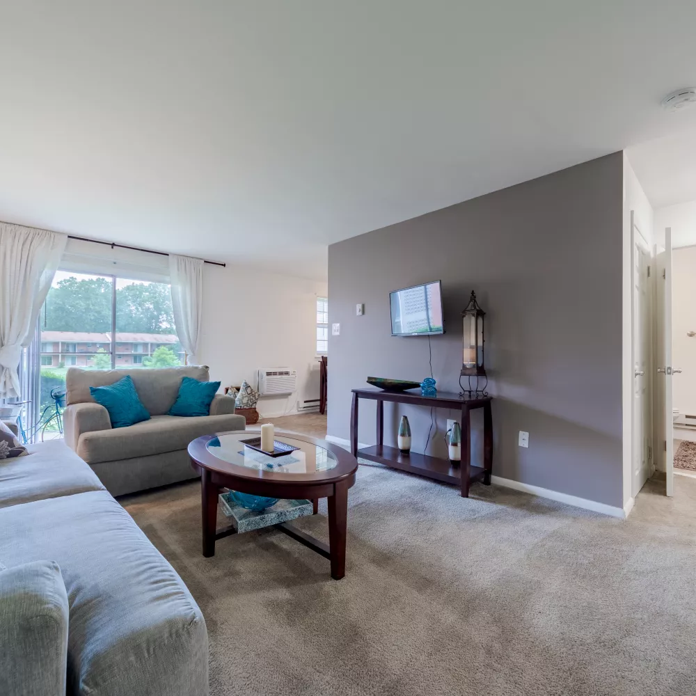 Cozy living room with light sofas, coffee table, and a wall-mounted TV; a bathroom is visible through an open doorway.