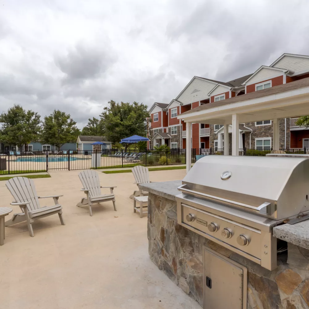 Outdoor grilling station with a large stainless steel grill on a stone counter, patio, pool, and apartment building under a cloudy sky.