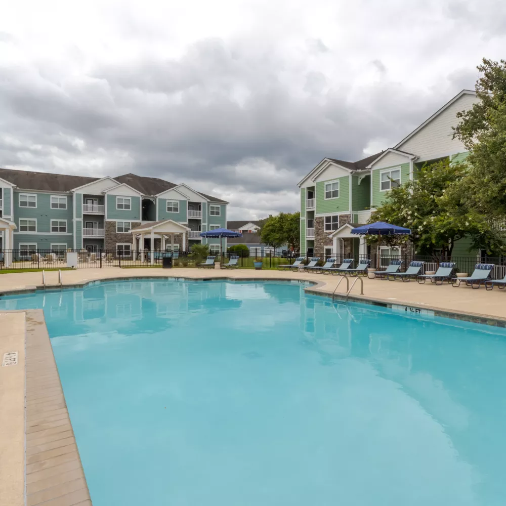 Outdoor swimming pool with lounge chairs, flanked by light blue and green apartment buildings under an overcast sky.