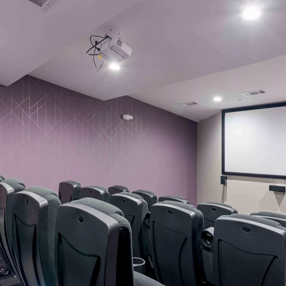Modern home theater with dark reclining seats, a large blank screen, and a ceiling-mounted projector. Purple geometric wall.