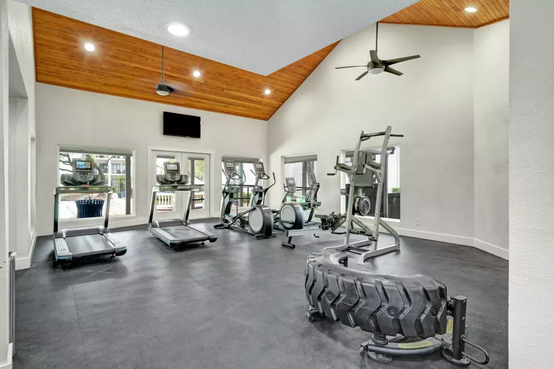 Spacious fitness center featuring a variety of modern exercise equipment, including treadmills, ellipticals, and weight machines. The room is bright and airy, with high ceilings, large mirrors, and a stylish wood-accented ceiling design.