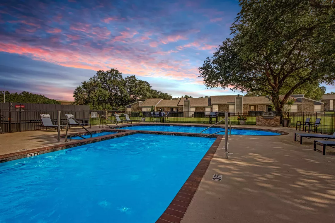 An outdoor pool illuminated at dusk, surrounded by lounge chairs, trees, and residential buildings under a vibrant sunset sky.