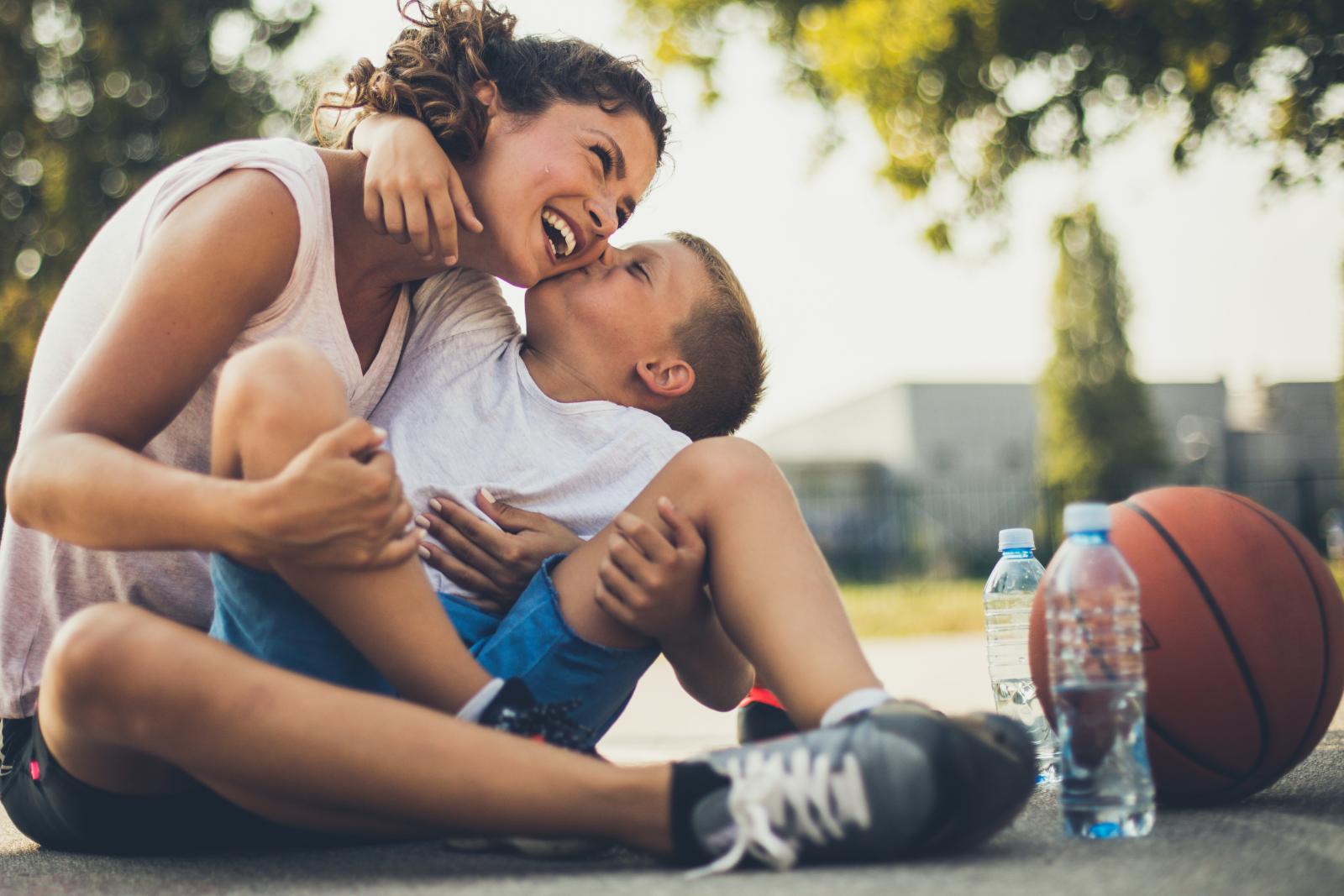A mother and her young son share a joyful moment, laughing and hugging while sitting on a basketball court with a basketball and water bottles nearby.