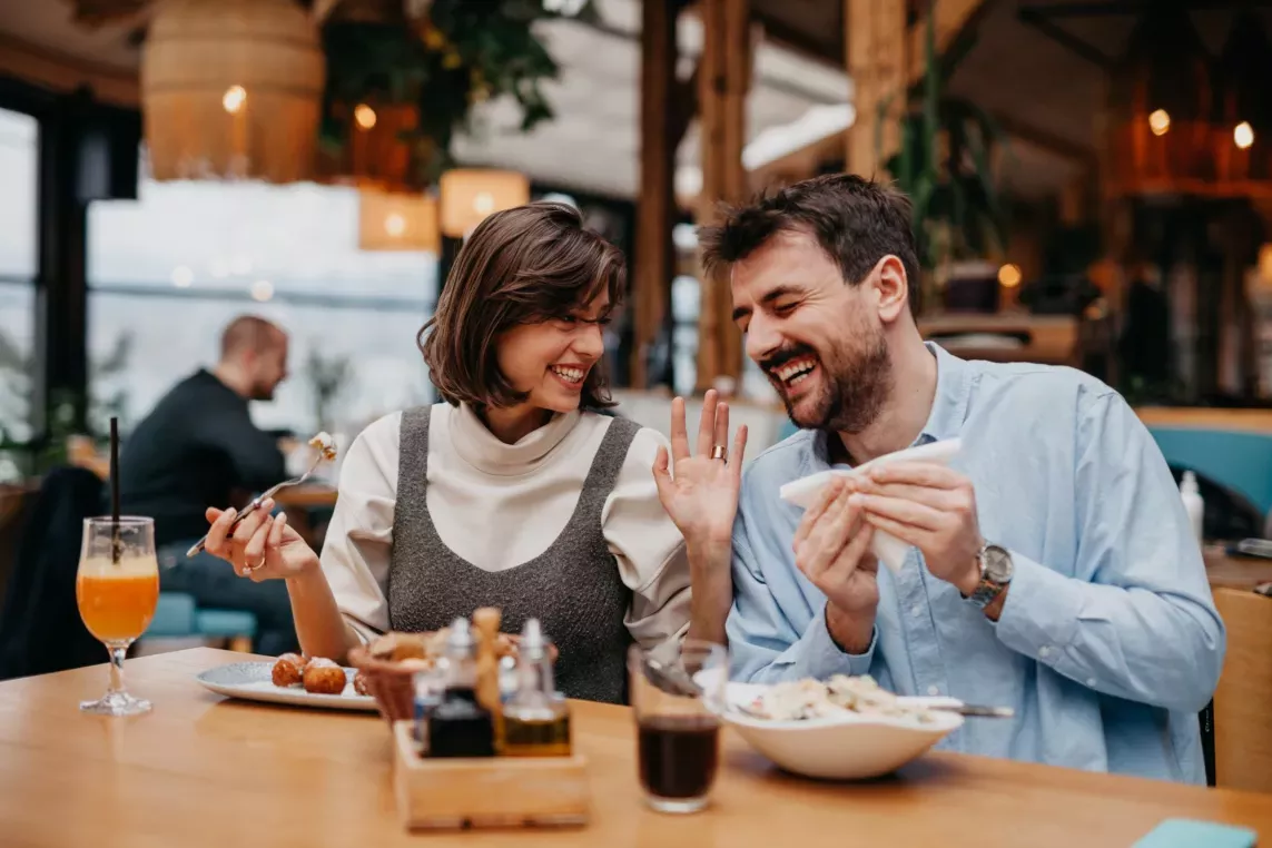  A smiling couple enjoying a meal together at a cozy restaurant, sharing food and laughter. The warm lighting and inviting atmosphere highlight a fun and relaxed dining experience.
