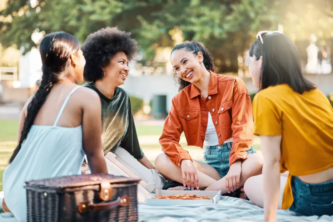  A group of friends enjoying a picnic outdoors on a sunny day, sitting on a blanket and sharing smiles and laughter. The setting highlights a relaxed, joyful atmosphere surrounded by greenery.