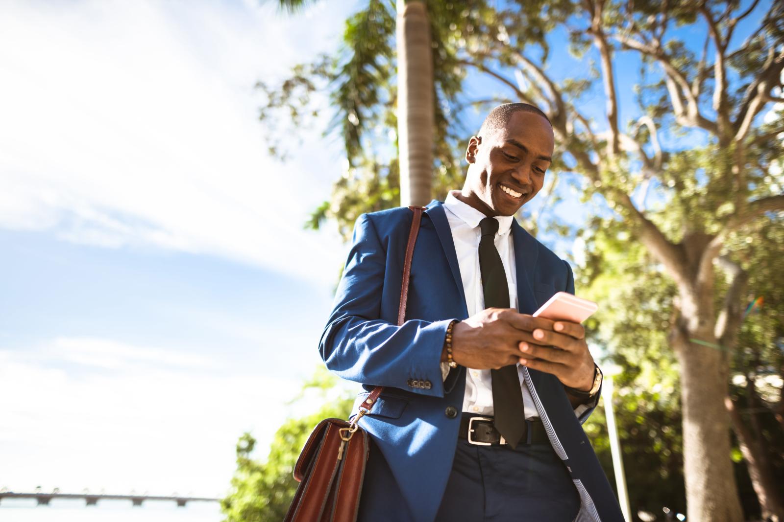 A businessman dressed in a blue suit smiles while looking at his phone outdoors, surrounded by trees and bright sunlight.