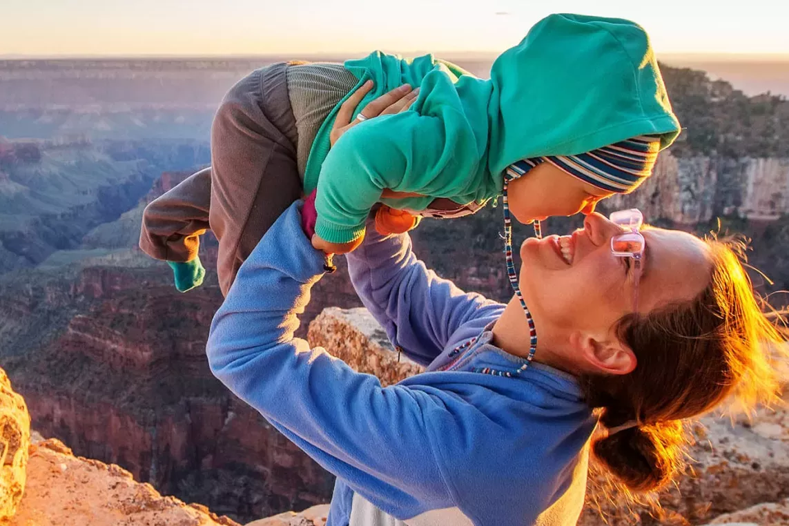 A woman playfully lifts a bundled-up child while sitting on a rocky ledge, with a breathtaking canyon landscape and sunset in the background.