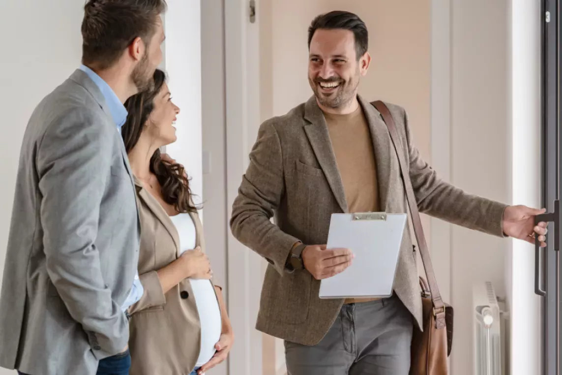 A real estate agent smiling while opening a door for a couple, with the woman appearing pregnant, during a property showing.