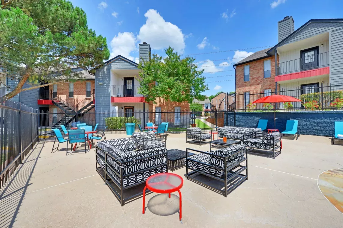 Vibrant outdoor community space featuring stylish seating areas with modern patio furniture, surrounded by colorful apartment buildings and lush greenery. The bright blue sky and open layout create an inviting and lively atmosphere for social gatherings.