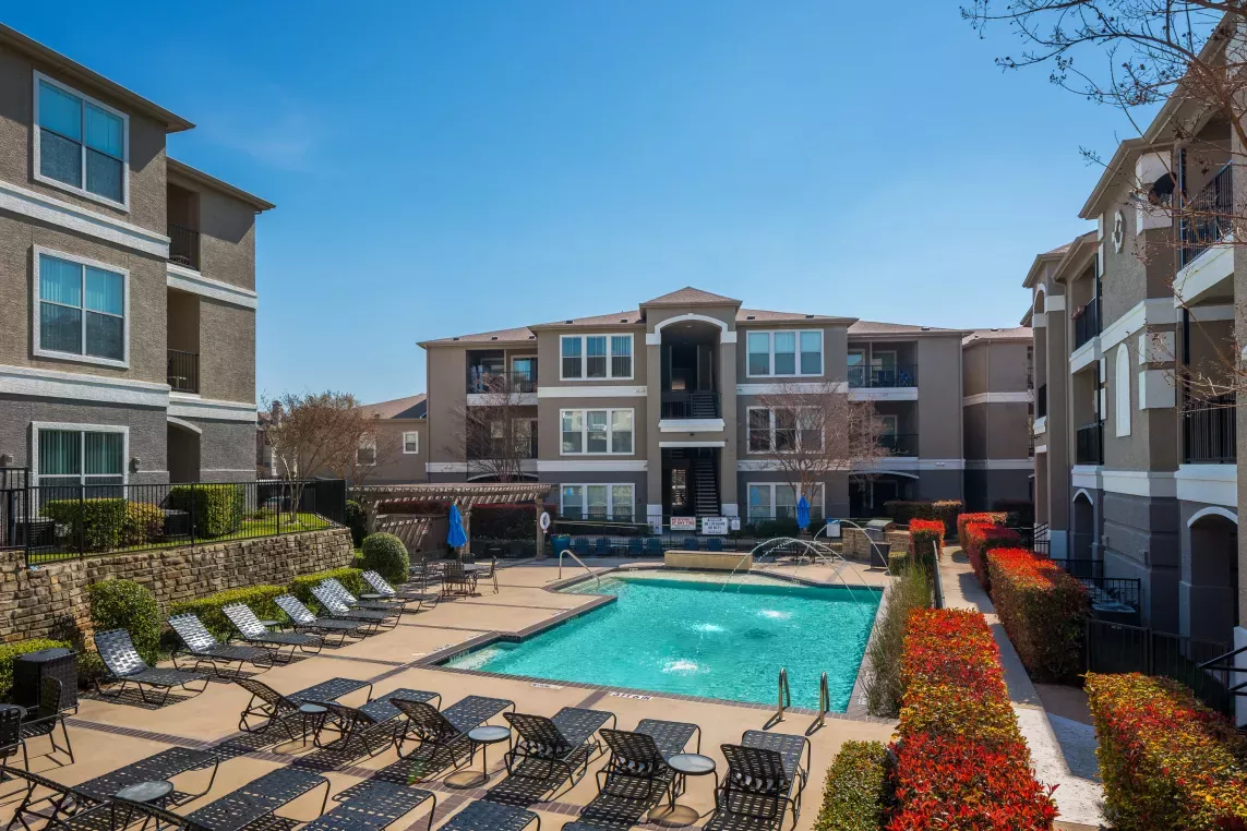 A bright courtyard with a sparkling pool, lined with lounge chairs, red shrubs, and modern apartment buildings in the background.