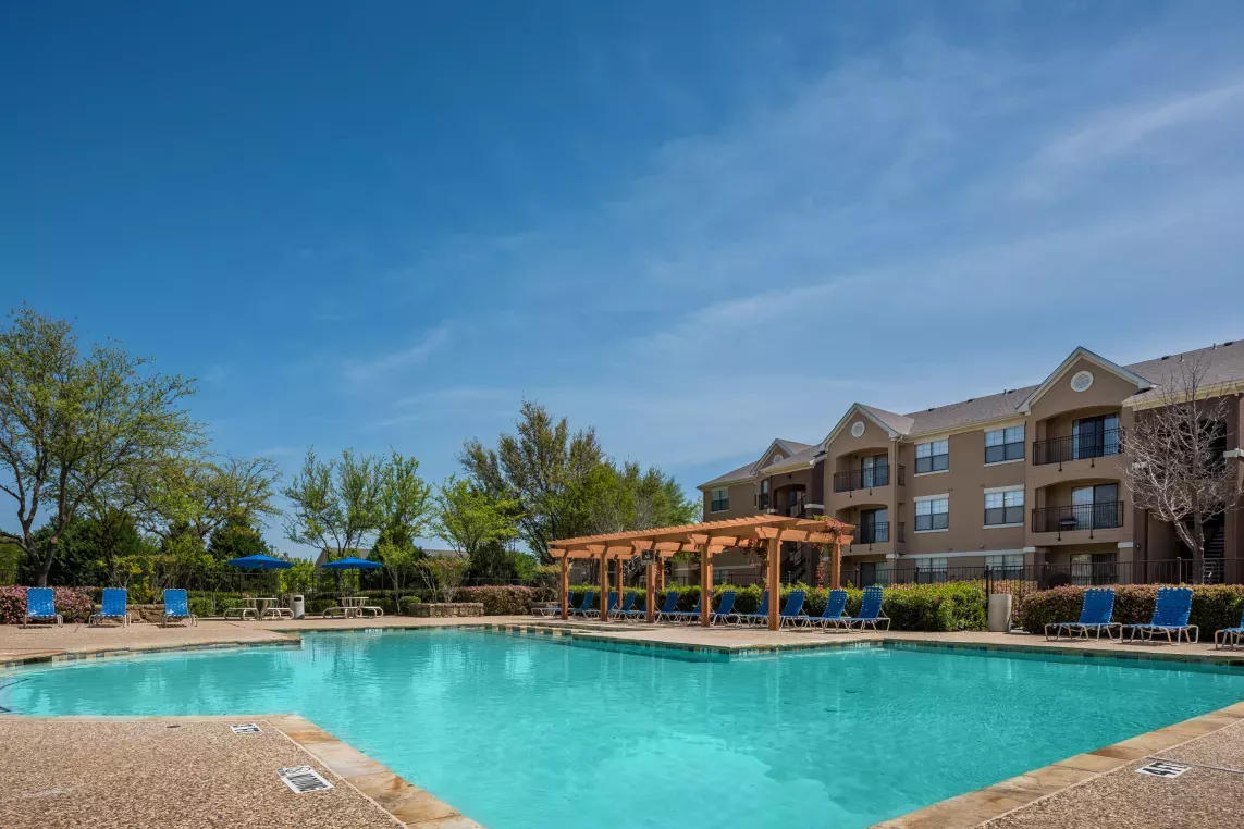 A sparkling outdoor pool surrounded by lounge chairs and a pergola, with apartment buildings and lush greenery in the background under a clear blue sky.