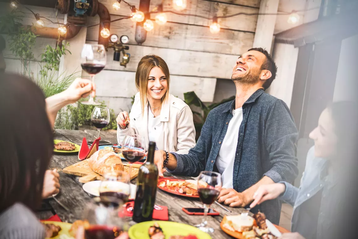 A group of friends enjoying food and wine at an outdoor table decorated with string lights and surrounded by rustic decor.