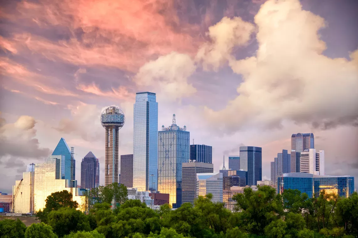 A city skyline with modern buildings and a prominent spherical structure, set against a dramatic sunset sky with lush greenery in the foreground.