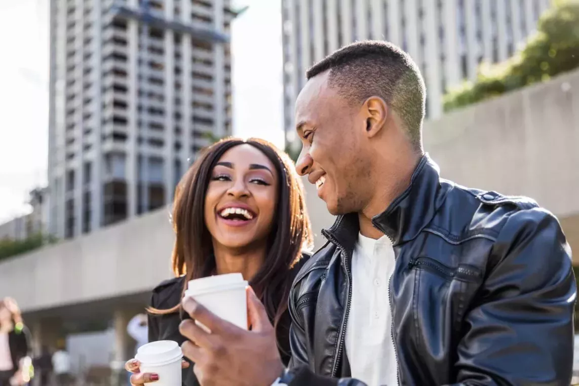 A man and woman are outdoors on a city street, laughing together. Both are smiling and appear to be enjoying a light-hearted moment while interacting with each other. The woman is holding a cellphone. They are dressed casually, with buildings in the background.