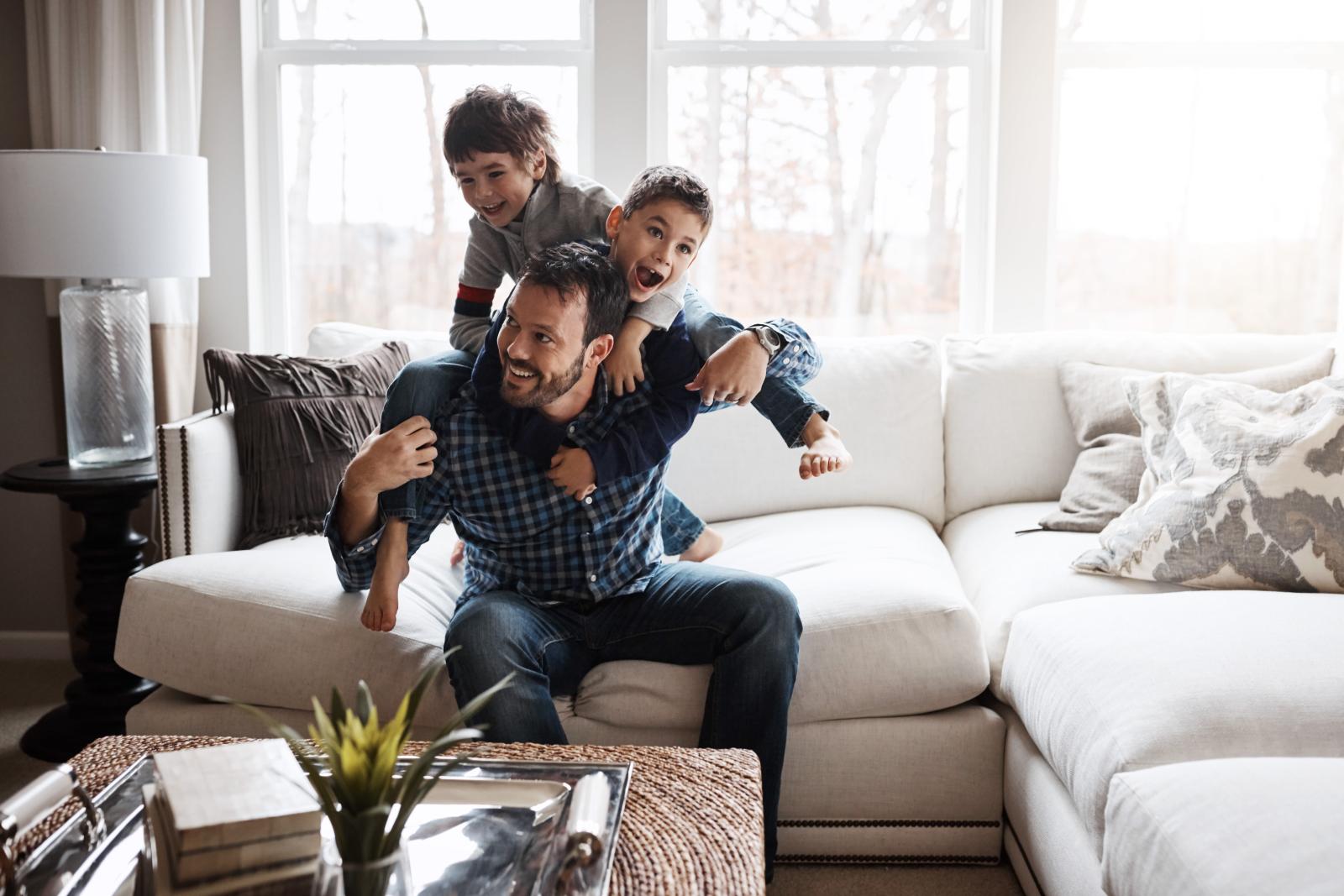 A father sitting on a cozy white couch, laughing and playing with his two young sons as they climb on his back in a bright living room.