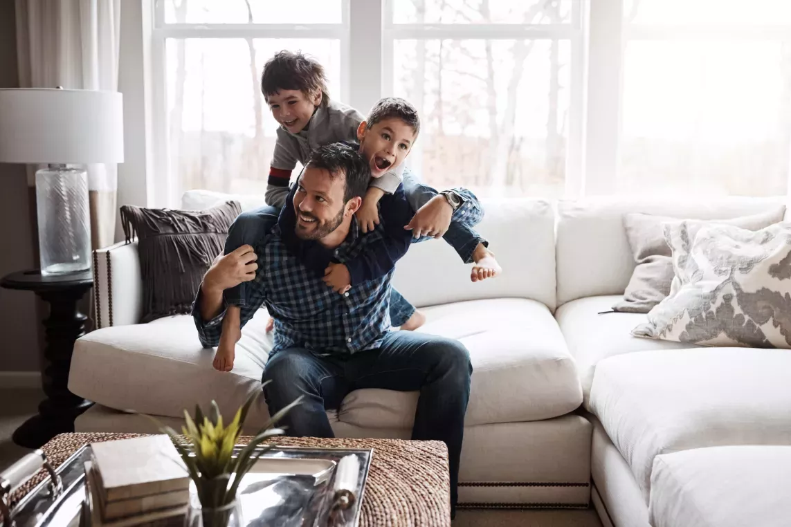 A joyful father enjoying quality time with his two sons, giving them piggyback rides in a cozy living room. The trio shares a fun and laughter-filled moment, creating lasting memories in a warm, comfortable home environment.