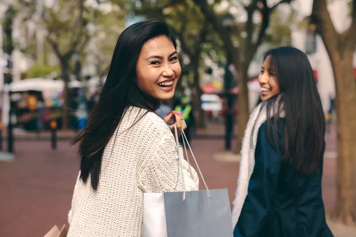 Two smiling friends carrying shopping bags while walking outdoors in an urban setting.
