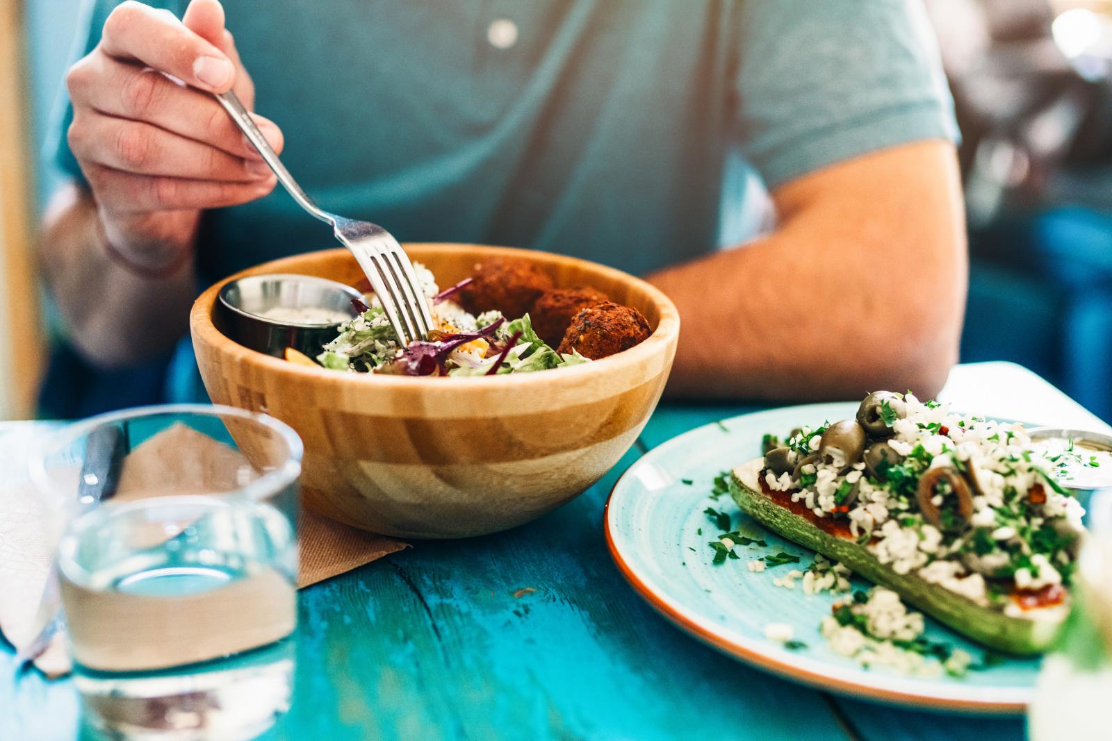 A person seated at a table, holding a fork and enjoying a fresh salad in a wooden bowl, alongside a plate of stuffed zucchini topped with olives and herbs.
