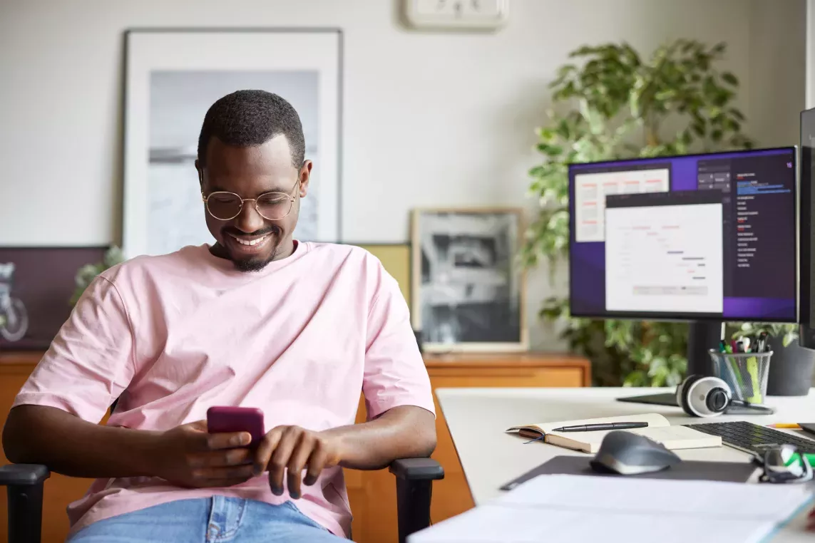A man sitting at a desk smiles while looking at his phone, surrounded by a computer monitor, notebook, and office decor.