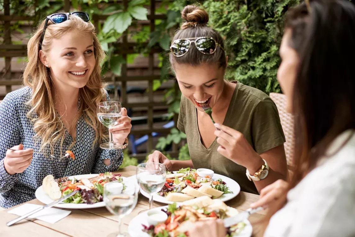 A group of three women enjoying a lunch together outdoors, sharing laughs as they eat fresh salads and drink water in a sunny, leafy setting. The relaxed atmosphere captures the joy of good food and even better company.