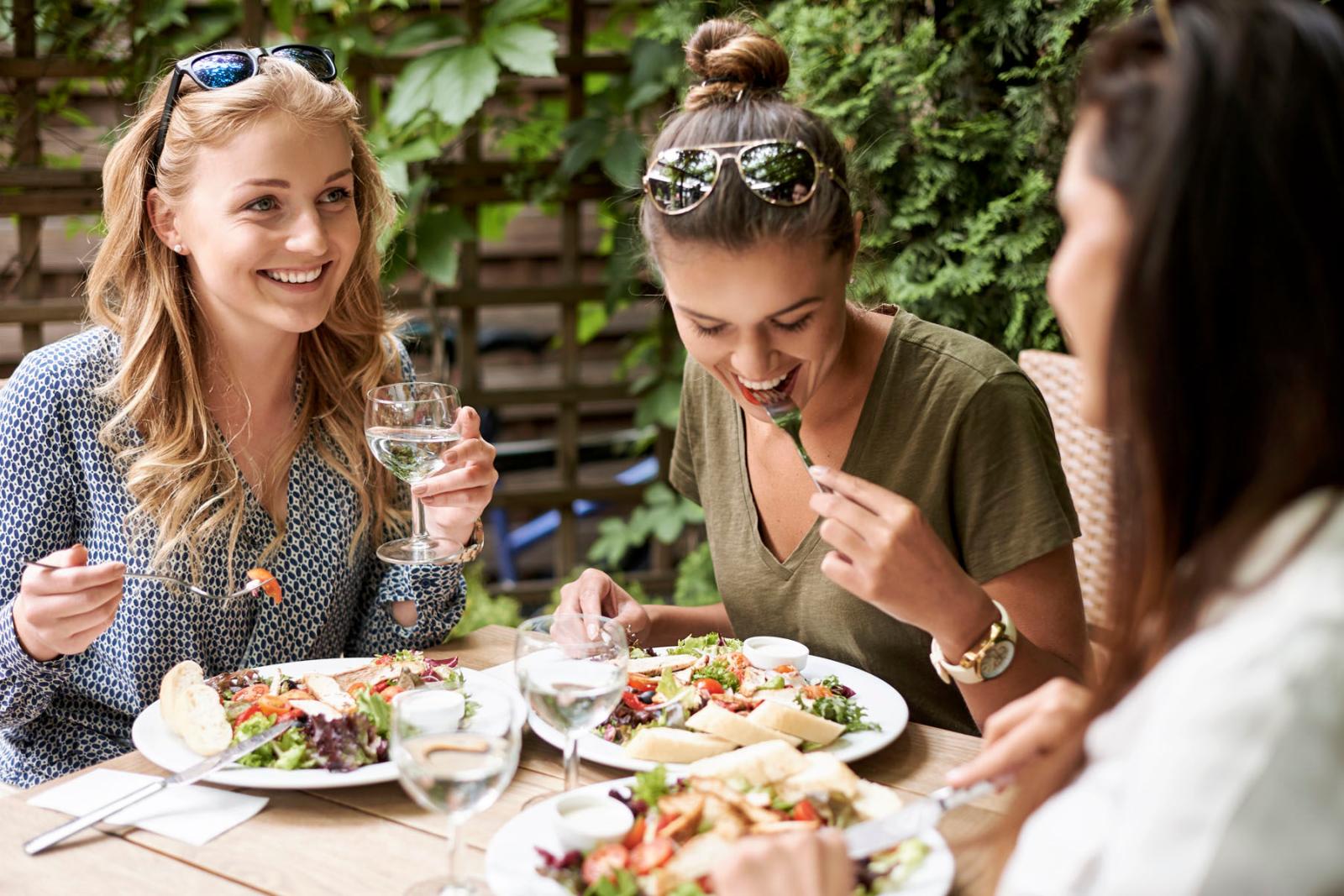 A group of three women enjoying a lunch together outdoors, sharing laughs as they eat fresh salads and drink water in a sunny, leafy setting. The relaxed atmosphere captures the joy of good food and even better company.
