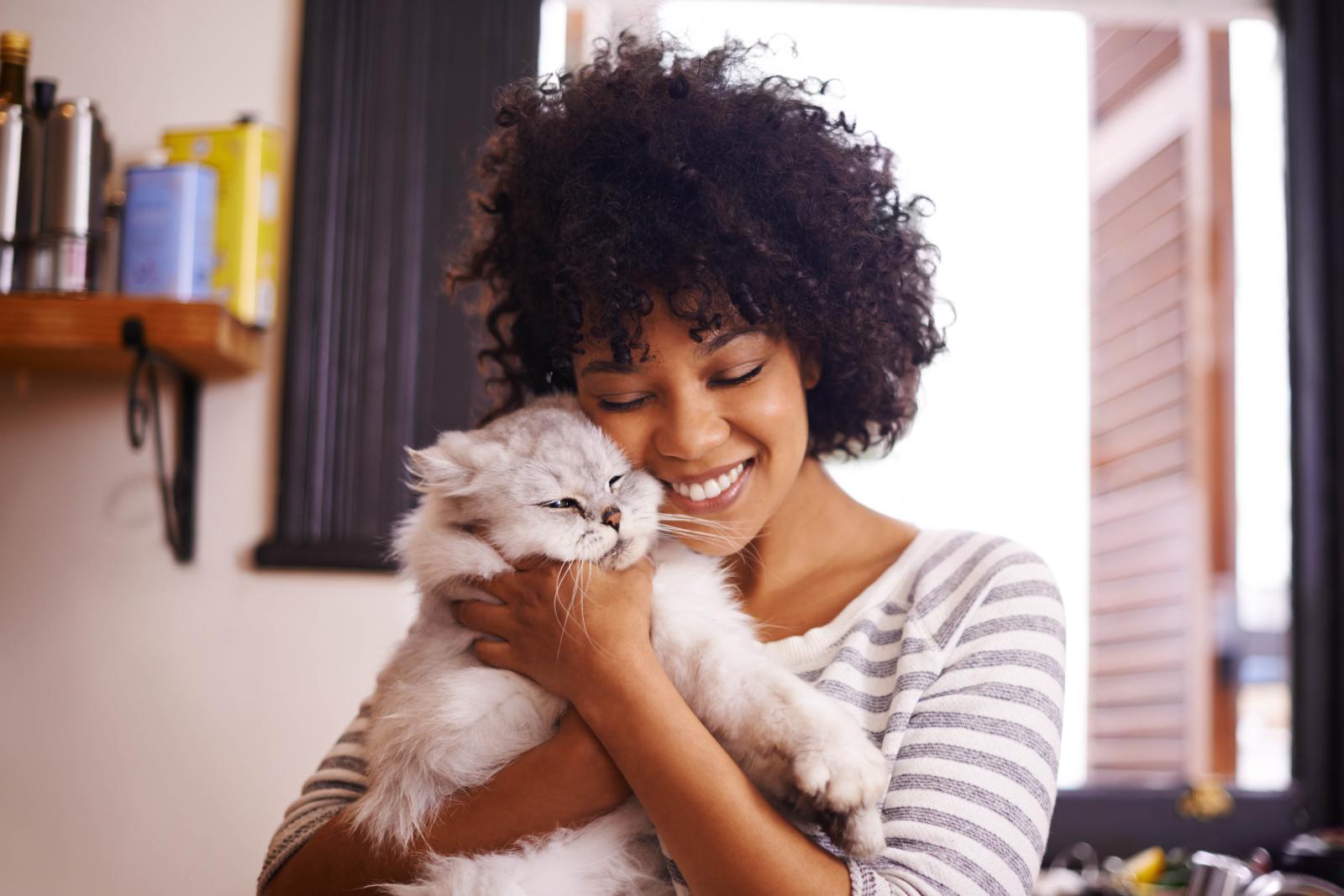A smiling woman cuddles a fluffy gray cat in a cozy kitchen setting.