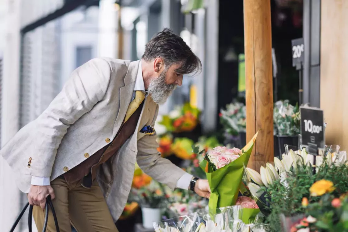 A stylish man with a gray beard and a light blazer selects a bouquet of pink roses at an outdoor flower market.