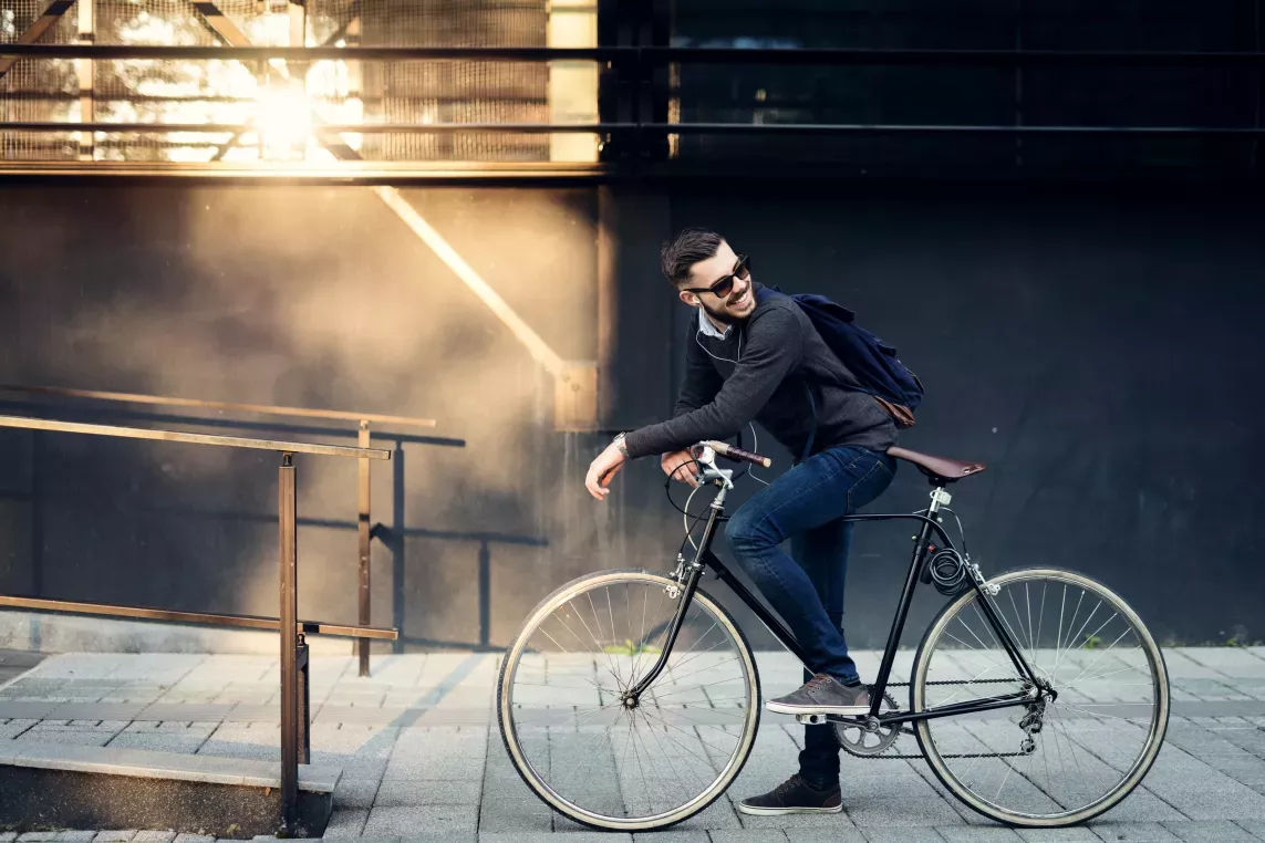 A young man wearing sunglasses and a backpack sits on a bicycle, smiling and enjoying the sunlight against an urban backdrop.