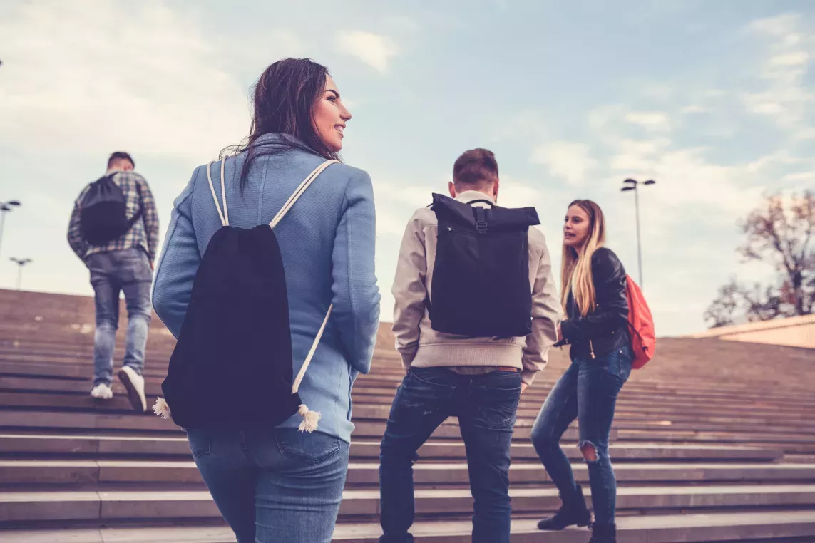 A group of young friends with backpacks walking up outdoor steps under a bright, partly cloudy sky.