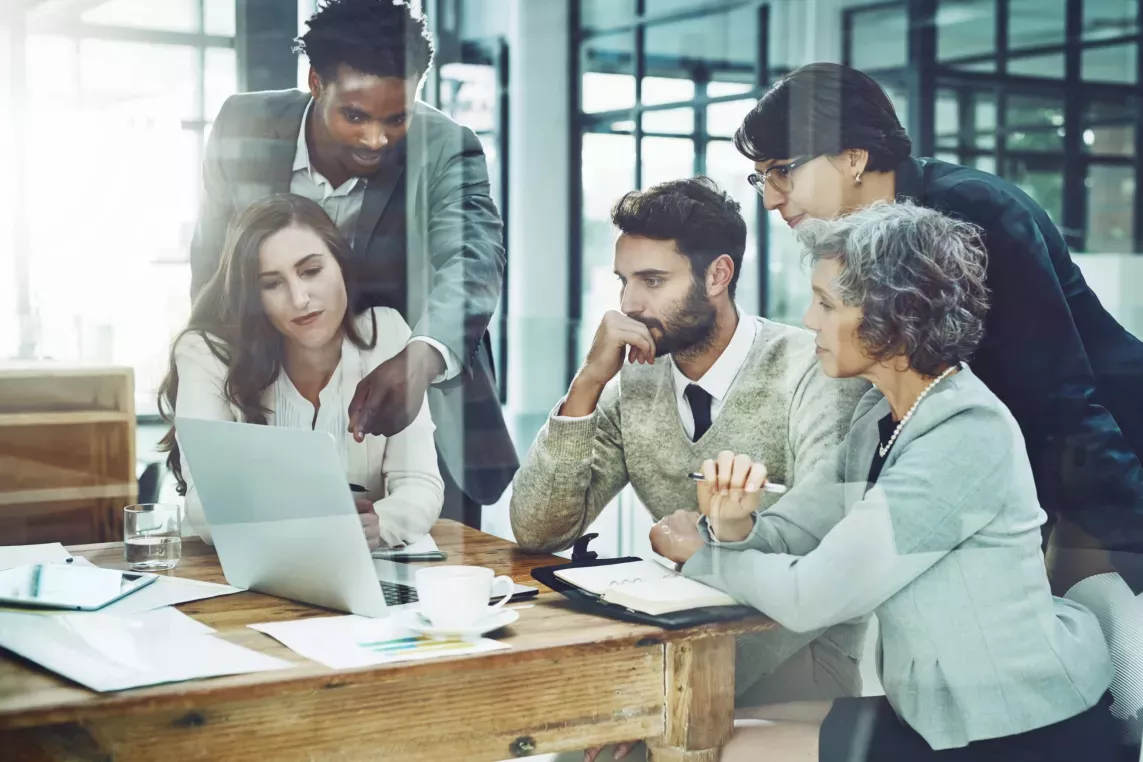 A diverse group of five professionals collaborates around a wooden table in a bright, modern office, focusing intently on a laptop screen during a discussion.