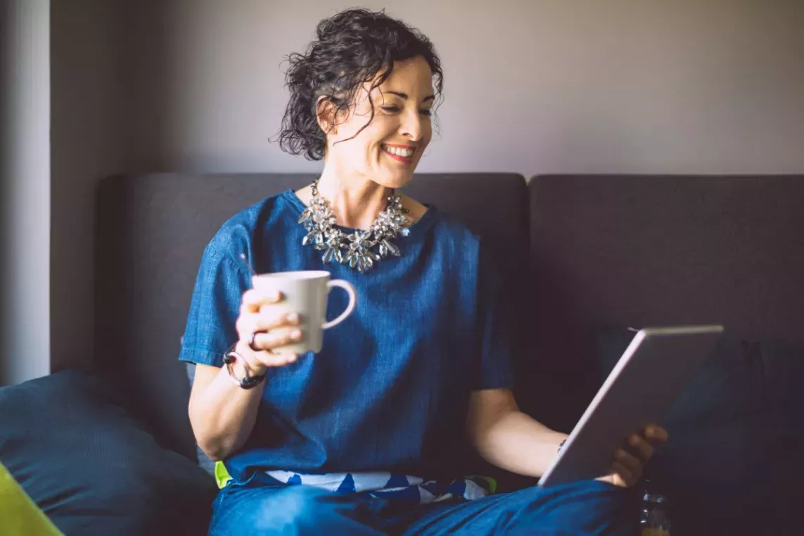 A smiling woman relaxes on a sofa, holding a cup of coffee and looking at a tablet.