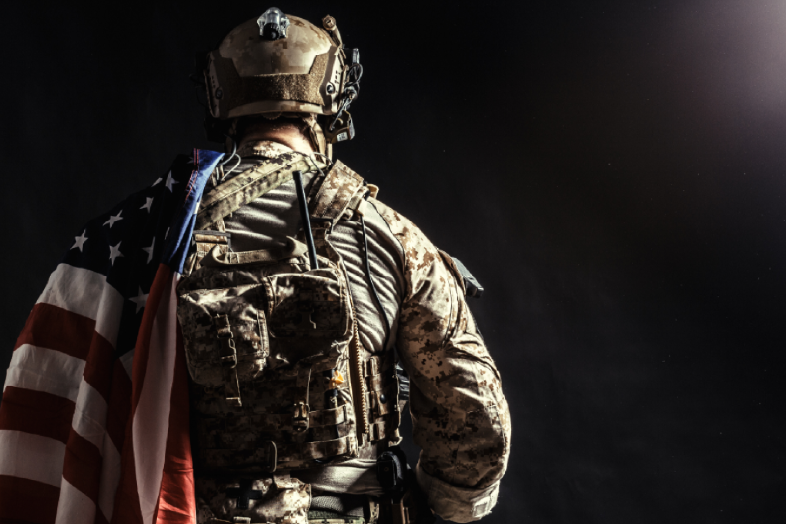 A soldier wearing tactical gear with an American flag draped over their shoulder, standing in a dimly lit setting.