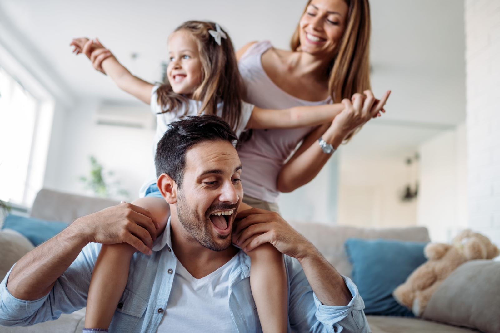 A family happily playing together at home, with the father carrying the child on his shoulders while the mother smiles alongside them.