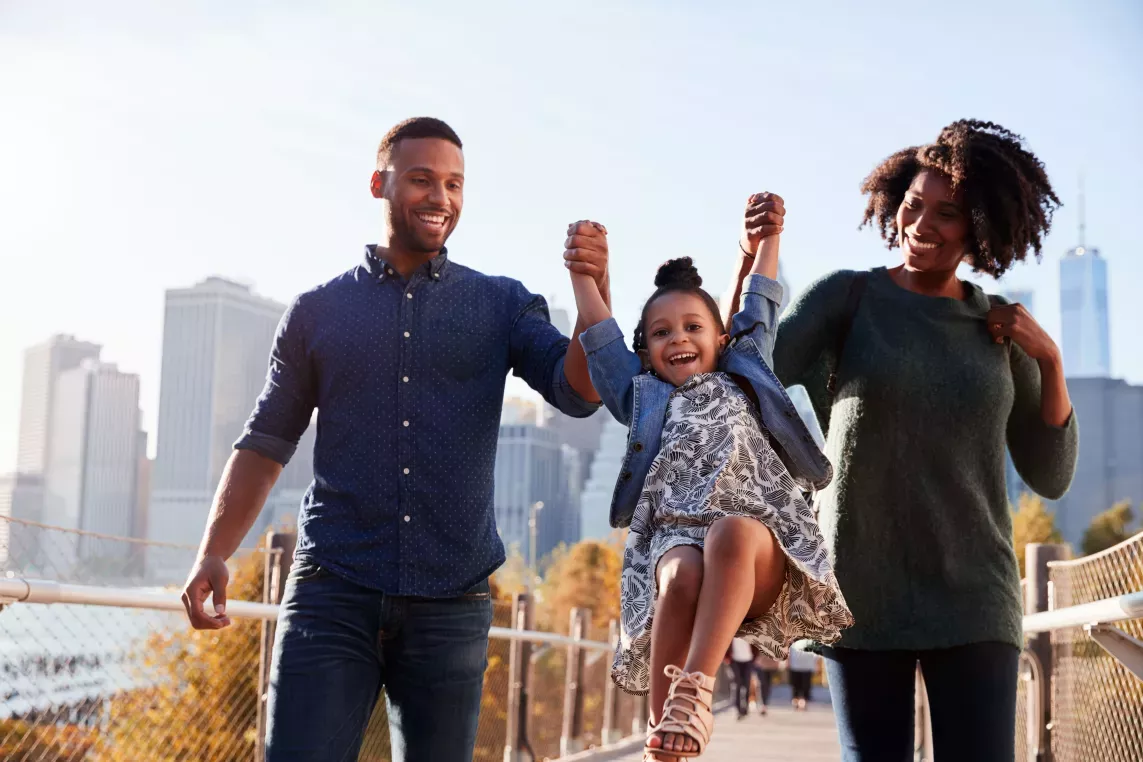 A joyful family walking on a boardwalk in the city, lifting their smiling child between them as skyscrapers loom in the background.