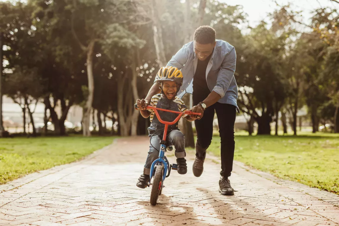 A joyful child rides a small red and blue bicycle with training wheels as a smiling adult helps guide and encourage them in a sunny park.