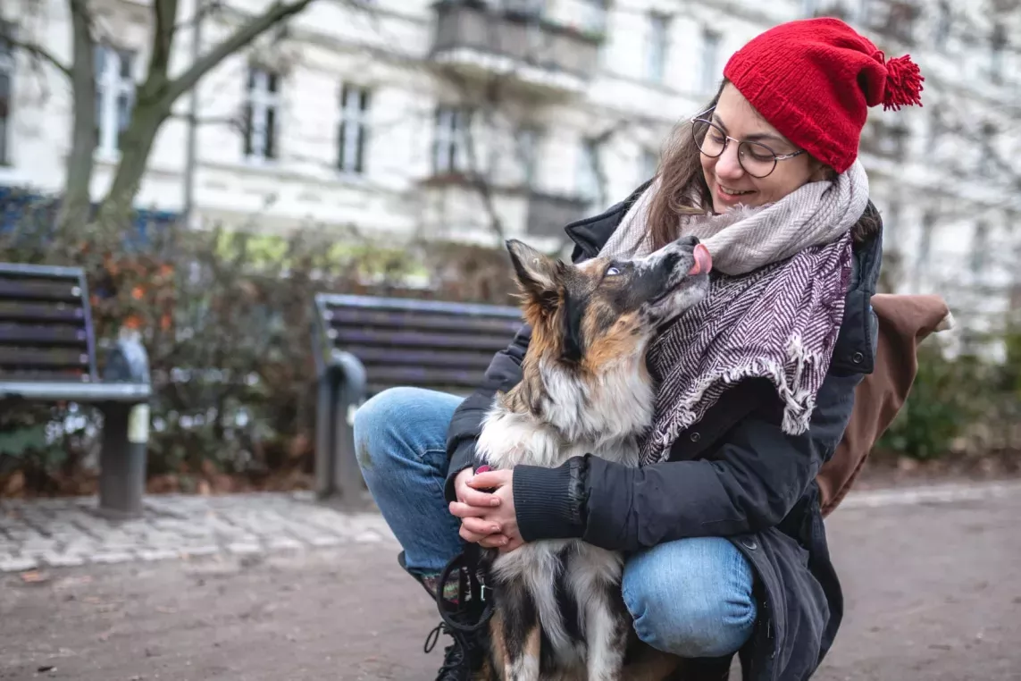 A woman wearing a red beanie kneels to embrace her dog in a park, both sharing a joyful moment.