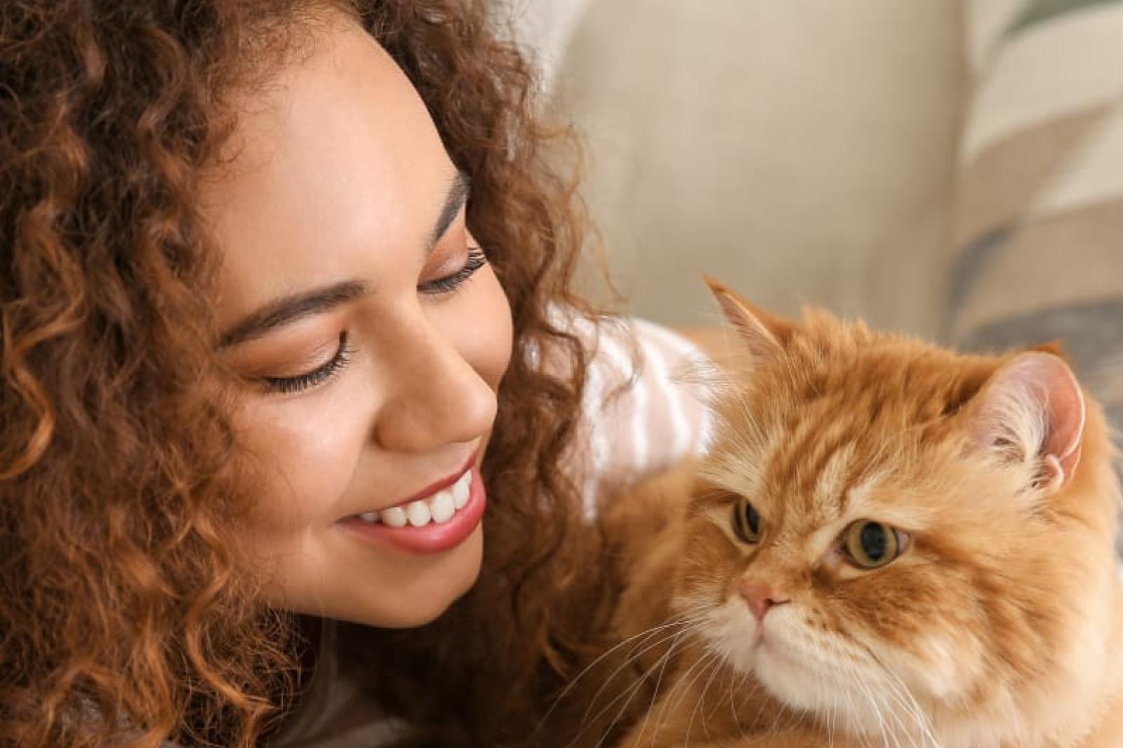 Woman smiling while lying down next to her fluffy orange cat, enjoying a peaceful moment together with a joyful and affectionate expression.