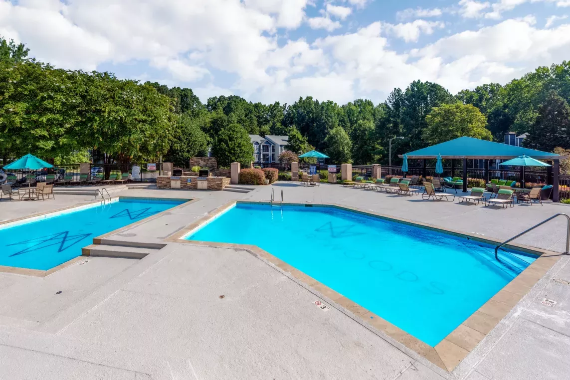 An inviting outdoor pool area with two sparkling blue pools surrounded by lounge chairs, umbrellas, and lush greenery in a serene residential setting.