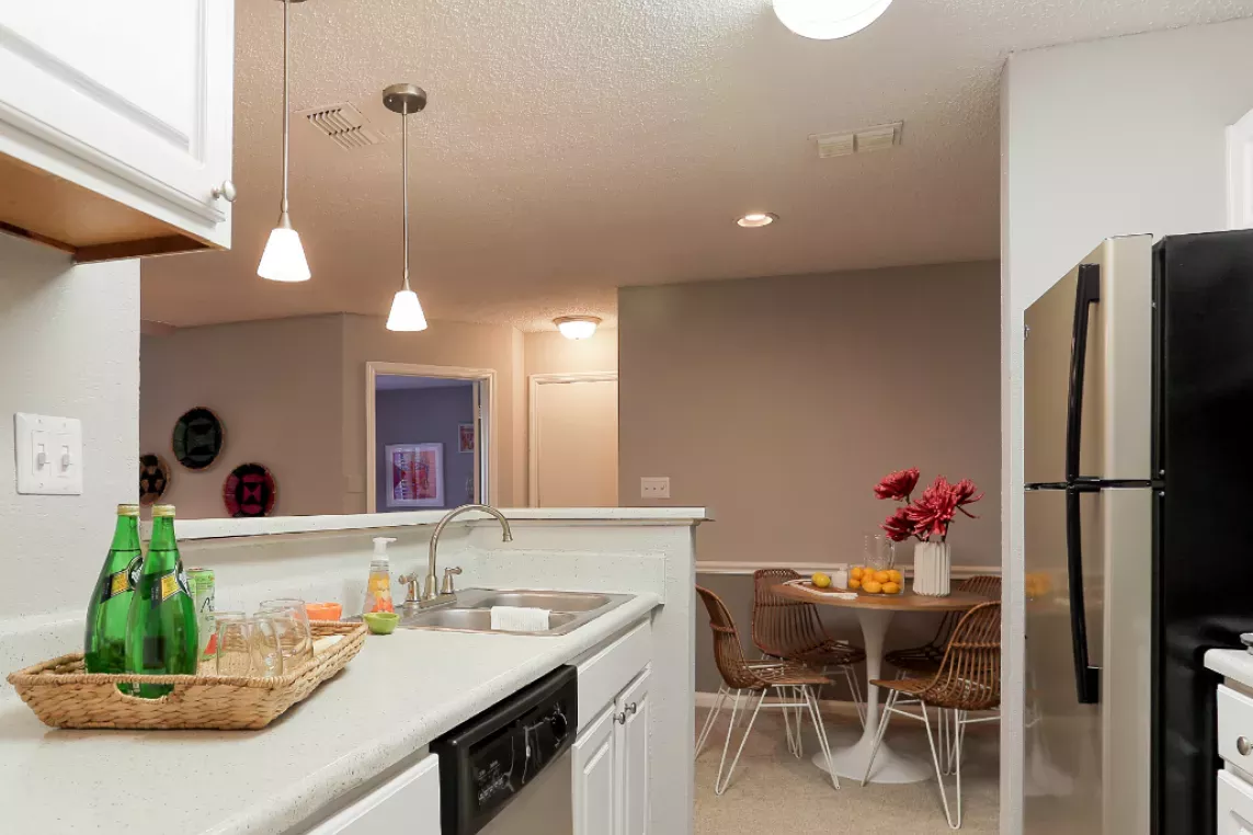 A modern kitchen setup featuring white cabinets, pendant lights, and a cozy dining area with wicker chairs and a table adorned with flowers and fruits.