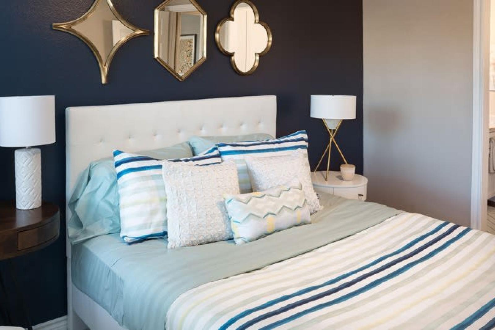 A stylish bedroom with a navy accent wall, decorative mirrors, a tufted white headboard, striped bedding, and modern side tables with lamps.